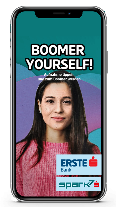 Darstellung eines Handys mit dem Facefilter "Boomer Yourself". Auf dem Bild sieht man eine Frau, die mit dem Filter gealtert wurde mit den Worten "Boomer Yourself! Aufnahme tippen und zum Boomer werden" und dem Erste Bank und Spark7 Logo in der rechten unteren Ecke.